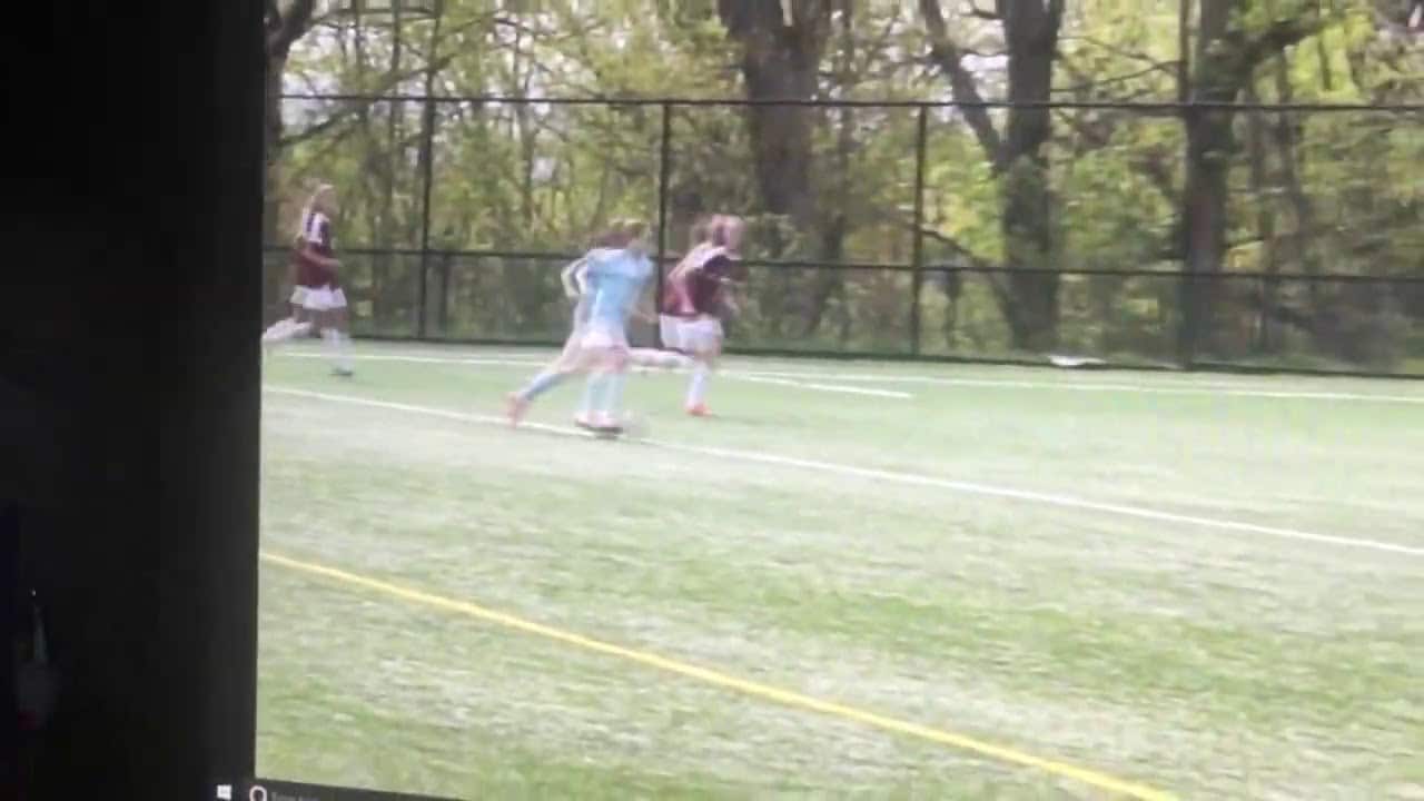 A soccer game with players on a green field, surrounded by trees and a fence. Players are in motion, with one in light blue and others in maroon and white.