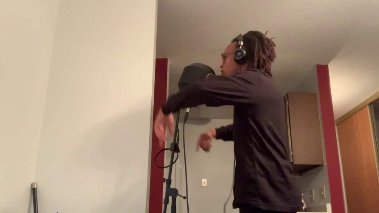 Person wearing headphones standing in front of a microphone in a room, gesturing with hands.