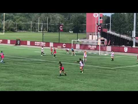 A women's soccer match on a green field with a stadium in the background. One team wears red and black, the other in white. The ball is near the goal defended by the white team's goalkeeper.