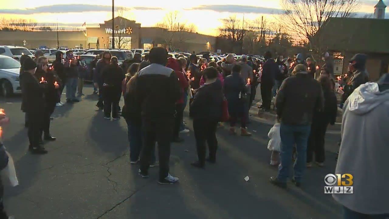 A group of people holding candles gathers in a parking lot at sunset, with a Walmart building visible in the background.