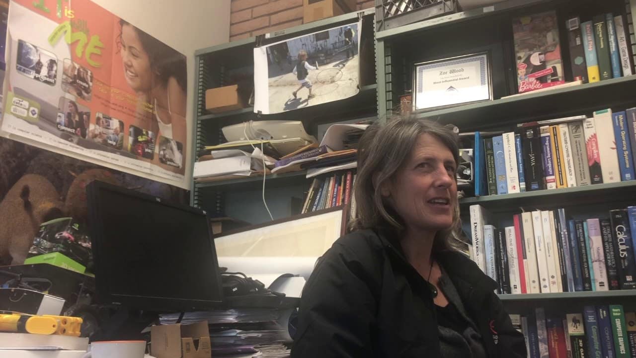 A woman sits in an office filled with books and posters, talking. Shelves are cluttered with various books and items.