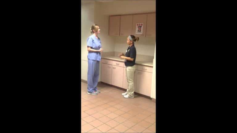 Two women, one in scrubs and one in casual work attire, stand and talk in a room with pink cabinets and tiled floor.