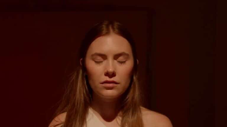 A woman with long brown hair stands with her eyes closed, facing forward, in soft, warm lighting against a dark background.